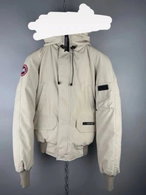 Canada goose  - Stilren beige bomberjacka från Canada Goose med huva och ribbade muddar. Jackan har flera fickor, dragkedja och snörning vid halsen. Klassisk patch på ärmen och diskret logotyp på fickan. Perfekt för kyliga dagar.