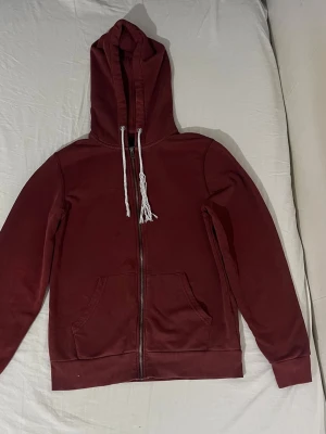 Vinröd hoodie med dragkedja - Säljer en vinröd hoodie med dragkedja och vita dragsnören. Hoodien har en klassisk känguruficka framtill och ribbade muddar vid ärmar och nederkant. Materialet är skönt, perfekt för chill dagar. Märke saknas. Den har fläckar ( se bilder). 
