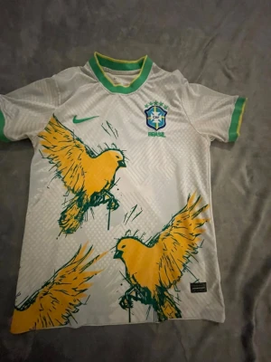 Vit Brasilien Nike fotbollströja - Snygg vit fotbollströja från Nike med Brasilien-emblem på bröstet. Tröjan har gröna detaljer vid krage och ärmslut samt stora gula fåglar tryckta på framsidan. Perfekt för dig som älskar fotboll och vill sticka ut på planen.