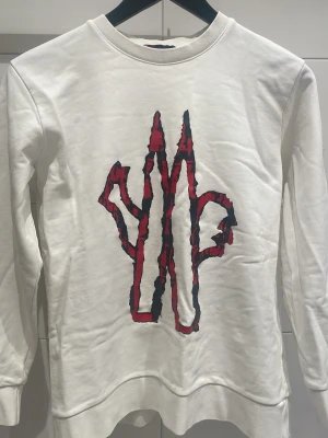 Vit sweatshirt från Moncler - Vit sweatshirt från Moncler med stort rött och svart grafiskt tryck på bröstet. Tröjan har rund halsringning och lång ärm med Moncler-logga på ärmen. Perfekt för en trendig och avslappnad stil.