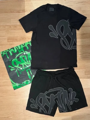 Svart set med graffiti-tryck - Säljer ett svart set med t-shirt och shorts, båda med stort grått graffiti-inspirerat tryck framtill. Setet har en avslappnad passform och är gjort i mjukt material, perfekt för en streetwear-look. Medföljer en matchande förvaringspåse i svart och grönt.