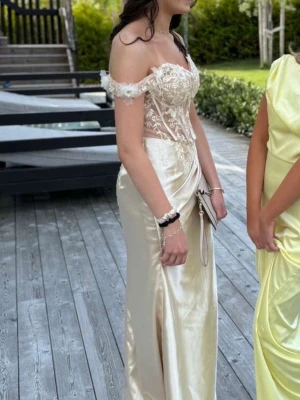 Champagnefärgad långklänning med spets - Elegant offshoulder långklänning i champagnefärgad satin med spetsdetaljer upptill och markerad midja. Klänningen har en figurnära passform, slits framtill och vackra volangdetaljer vid axlarna. Perfekt för bal eller student.