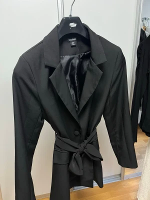 Svart kavaj med knytband Boohoo - Stilren svart kavaj från Boohoo med klassisk krage, knytband i midjan och två fickor framtill. Kavajen har en knappstängning och är helfodrad med blankt tyg på insidan. Perfekt för en trendig och snygg look.