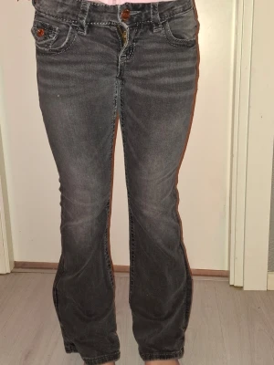 Grå bootcut jeans med broderade fickor - Säljer ett par grå bootcut jeans med snygga slitningar och broderade bakfickor. Jeansen har bruna knappar och detaljer, samt klassisk femficksmodell. Perfekta för dig som gillar en avslappnad men trendig look.