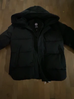 Svart dunjacka från Canada Goose - Säljer en svart dunjacka från Canada Goose med huva och klassisk patch på ärmen. Jackan har en puffig design, stora fickor framtill och dragkedja med vindslå. Perfekt för kalla vinterdagar och riktigt varm.