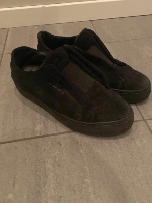 Svarta sneakers från Axel Arigato - Stilrena svarta sneakers från Axel Arigato i mocka med minimalistisk design och diskret logga i guld på sidan. Skorna har låg profil och elastiska band istället för klassiska skosnören för en clean look.