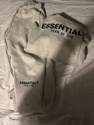 Grå mjukisbyxor från Essentials - Säljer ett par ljusgrå mjukisbyxor från Fear of God Essentials med svart logga tryckt på benet. Byxorna har resår i midjan och vid bensluten för en skön och avslappnad passform. Perfekta för chill dagar och streetwear-stil.