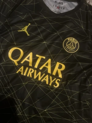 Svart PSG matchtröja Jordan x Nike - Snygg svart PSG matchtröja med gula detaljer och grafiskt mönster. Tröjan har Paris Saint-Germain-logga, Jordan-logga och Qatar Airways-tryck på bröstet samt GOAT-tryck på ärmen. Tillverkad i lätt och ventilerande material, perfekt för fotboll eller träning.