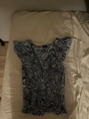 Blommig blå och vit blus - Söt blus med blått och vitt blommönster, v-ringad och med korta volangärmar. Smockad midja och lätt volangkant nedtill ger en feminin look. Perfekt för dig som gillar romantiska detaljer och mjuka material.