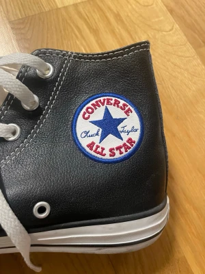 Svarta Converse All Star high-top - Klassiska Converse All Star high-top sneakers i svart skinn med vita kontrastsömmar och vita snören. Ikonisk rund logga på sidan och vit tåhätta i gummi. Sulan är brun undertill och skon har metallöljetter för snörningen. Perfekta för en cool och tidlös stil. Strl 38 Köpti i USA. US 5.5, svensk strlk 38