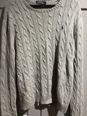 Grå kabelstickad tröja Polo Ralph Lauren - Kabelstickad grå tröja från Polo Ralph Lauren med rund halsringning och diskret broderad logga på bröstet. Tröjan har långa ärmar och ribbade muddar vid ärmslut och nederkant. Perfekt för en stilren och klassisk look. Passar L-XL. Priset kan diskuteras