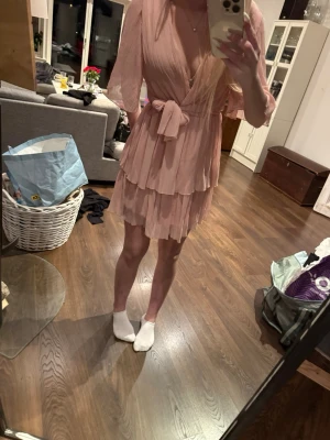 Rosa omlottklänning med volanger - Söt rosa kort omlottklänning med volanger och knyt i midjan. Klänningen har v-ringning och korta, fladdriga ärmar. Perfekt för dig som gillar en romantisk och feminin stil.