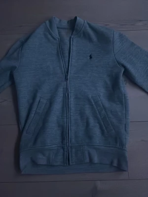 Grå ziptröja från Polo Ralph Lauren - Snygg grå ziptröja från Polo Ralph Lauren med klassisk broderad logga på bröstet. Tröjan har två fickor framtill, ribbade muddar och är tillverkad i mjukt material. Perfekt för en avslappnad och stilren look.