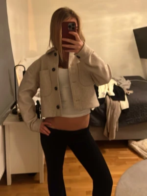 Beige cropped jacka med stora fickor - Snygg beige cropped jacka med stora framfickor och svarta knappar. Jackan har en boxig passform och är tillverkad i ett mjukt, filtliknande material. Perfekt för lager-på-lager och ger en trendig look till din outfit.