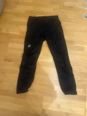 Svarta aktivitetsbyxor från RevolutionRace - Svarta byxor från RevolutionRace med smal passform och flera fickor. Byxorna har en diskret logga på ena benet och är tillverkade i ett slitstarkt material som passar perfekt för en aktiv livsstil. Tyvärr fungerar ibland inte dragkedjorna på insida lår utan nätet syns igenom. Säkert något som kan fixas av den händiga! I övrigt mycket fint skick
