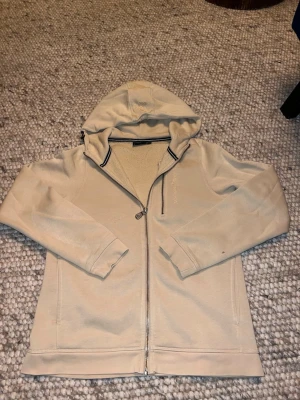 Beige hoodie från Sail Racing - Snygg beige hoodie från Sail Racing med dragkedja hela vägen och huva. Mjukt material och diskreta detaljer, perfekt för en chill och stilren look. Svarta och vita ränder på insidan av kragen ger en sportig touch. Passar dig som gillar klassisk streetwear.