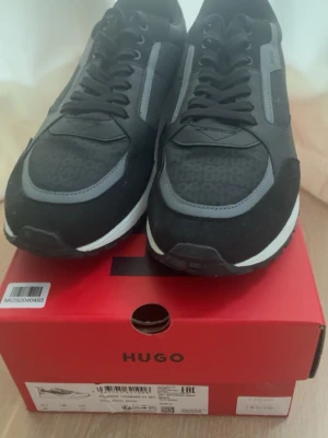 Svarta HUGO BOSS sneakers.  - Stilrena svarta sneakers från HUGO med grå detaljer och logotypmönster på tån. Skorna har en klassisk låg siluett, svarta skosnören och vit sula. Materialmix av mocka och textil ger en modern look som passar till många outfits. Använda 2 gånger då dom var förstora. Nypris 2099kr från NK.