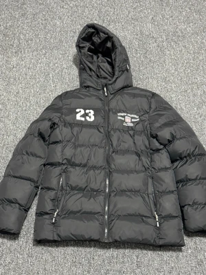 Svart dunjacka med huva och tryck - Svart dunjacka från Vingåker Denim med huva, dragkedja framtill och två fickor med dragkedja. Jackan har vitt tryck med siffran 23 och ett emblem på bröstet. Perfekt för kalla vinterdagar och har en sportig look.