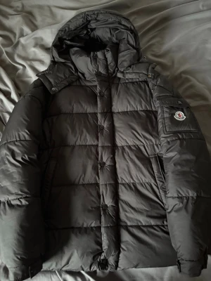 Svart dunjacka med huva från Moncler - Snygg svart dunjacka från Moncler med avtagbar huva och klassisk Moncler-logga på ärmen. Jackan har dragkedja och knappar framtill, två sidofickor och quiltad design. Perfekt för kalla vinterdagar och riktigt varm tack vare dunfyllningen.