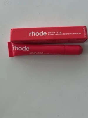 rhode Peptide Lip Tint röd läppglans - Säljer ett rhode Peptide Lip Tint i en snygg röd nyans. Läppglanset kommer i en tub med vit text och har en krämig formula som ger glans och färg till läpparna. Perfekt för dig som vill ha återfuktade och glossiga läppar med en hint av färg.