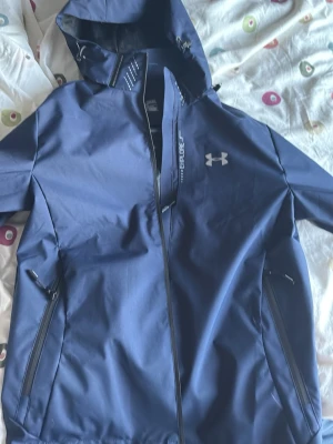 Blå vindjacka från Under Armour - Snygg blå vindjacka från Under Armour med huva och dragkedja framtill. Jackan har två dragkedjeförsedda fickor på sidorna och en diskret logga på bröstet. Perfekt för blåsiga dagar och sportiga outfits. Den är Vattentätt 