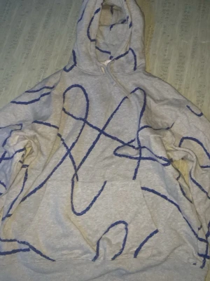 Grå hoodie med blått grafiskt mönster - Säljer en grå hoodie med blått grafiskt mönster över hela plagget. Hoodien har en stor huva och en klassisk magficka framtill. Tillverkad i mjukt sweatshirtmaterial, perfekt för en avslappnad streetwear-look.