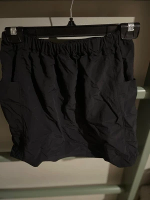 Svart kortkjol med resårmidja - En svart kortkjol med resår i midjan och praktiska sidofickor. Kjolen har en enkel och sportig look, perfekt för dig som gillar en avslappnad stil. Tillverkad i ett lätt syntetmaterial som känns bekvämt och smidigt.