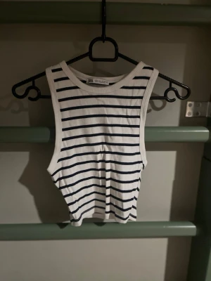 Randig ribbad linnetopp från Zara - Säljer en vit ribbad linnetopp från Zara med svarta horisontella ränder. Toppen är croppad och ärmlös med breda axelband och rund halsringning. Perfekt för en fräsch och avslappnad look. Dock väldigt kort 