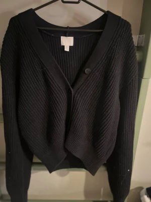 Svart ribbstickad kofta från H&M - Snygg svart ribbstickad kofta från H&M med v-ringning och knäppning framtill. Koftan har långa ärmar och en avslappnad passform, perfekt att slänga över en t-shirt eller topp. Klassisk och enkel design som funkar till det mesta.
