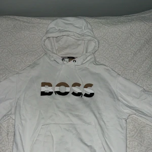 Vit oversized hoodie från BOSS - Snygg vit hoodie från BOSS med oversized fit och stor huva. Framtill står det 'BOSS' i stora bokstäver med brun, svart och vit färg. Hoodien har dragsko i huvan och en klassisk magficka. Perfekt för en chill och trendig look.
