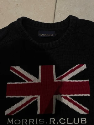 stickad tröja Morris R. Club - Marinblå stickad tröja från Morris R. Club med stor Union Jack-flagga i rött, vitt och grått på bröstet. Rund halsringning och ribbade muddar vid ärmslut och nederkant. Perfekt för dig som gillar klassisk brittisk stil.