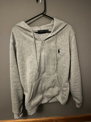Grå hoodie från Polo Ralph Lauren - Snygg grå hoodie med dragkedja från Polo Ralph Lauren. Klassisk design med huva, dragsnören och kängurufickor framtill. Broderad marinblå logga på bröstet. Perfekt för en avslappnad och stilren look.
