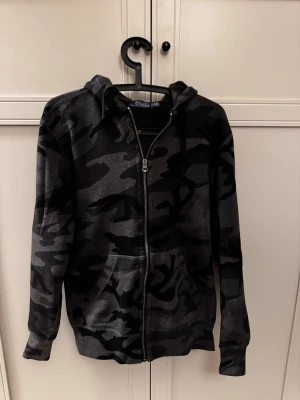 Camo hoodie Polo Ralph Lauren - Snygg hoodie med dragkedja från Polo Ralph Lauren i mörkgrått camomönster. Perfekt för en avslappnad streetstyle-look. Tillverkad i mjukt material för extra komfort.