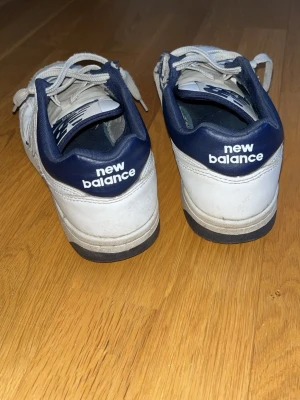 Vita New Balance sneakers med blå detaljer - Klassiska vita sneakers från New Balance med mörkblå detaljer och logga på sidan samt hälen. Skorna har lågt skaft, vita snören och en slitstark sula. Perfekta för dig som gillar retrostil och sportig vibe.
