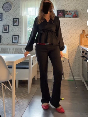 Svart omlottopp i satin - Snygg svart omlottopp i glansigt satinmaterial med lång ärm och volangdetalj i midjan. Toppen har en feminin v-ringning och en lös passform som ger en avslappnad men chic look. Från missay