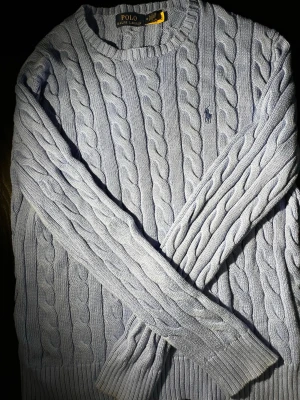 Ljusblå kabelstickad tröja Polo Ralph Lauren - Snygg ljusblå kabelstickad tröja från Polo Ralph Lauren med rund halsringning och diskret broderad logga på bröstet. Tröjan har långa ärmar och ribbade muddar vid ärmslut och nederkant. Perfekt för dig som gillar klassisk och stilren look. Tröjan är äkte och har en liten deffekt/hål som är lagat vid naken🙌