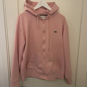 Ljusrosa hoodie från Lacoste - Säljer en ljusrosa hoodie från Lacoste i klassisk modell med dragkedja framtill, huva med snörning och känguruficka. Tröjan har det ikoniska krokodilmärket broderat på bröstet och är tillverkad i mjuk bomull. Perfekt för en chill och avslappnad stil.
