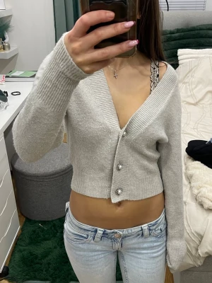 Ljusgrå cropped kofta med pärlknappar - Trendig ljusgrå cropped kofta med djup V-ringning och tre dekorativa pärlknappar framtill. Koftan har långa ärmar och breda ribbade muddar. Perfekt att styla med jeans eller kjol för en chill look.