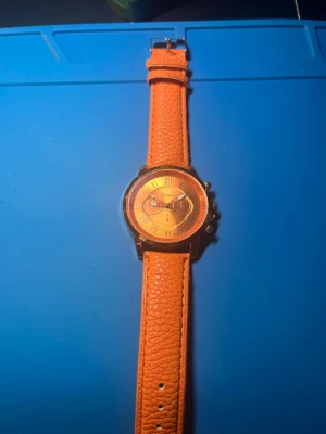 Orange klocka med läderband - Snygg klocka med orange urtavla och matchande orange band i läderimitation. Urtavlan har romerska siffror och flera små visare för extra detaljerad look. Perfekt accessoar för dig som vill sticka ut med färg. Behövs nya batterier 