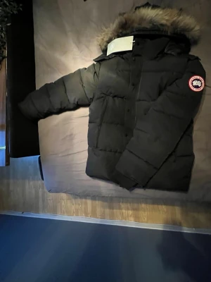 Svart dunjacka från Canada Goose - Säljer en svart dunjacka från Canada Goose med klassisk patch på ärmen och stor huva med fluffig pälsimitation. Jackan har en rak passform, dragkedja och knappar framtill samt flera praktiska fickor. Perfekt för kalla vinterdagar.