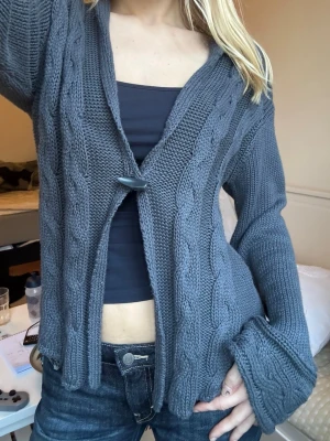 Mörkgrå kabelstickad kofta - Mysig mörkgrå kabelstickad kofta med en stor knapp framtill. Koftan har breda muddar, djup v-ringning och avslappnad passform. Perfekt att slänga över en topp för en chill look. 60% bomull