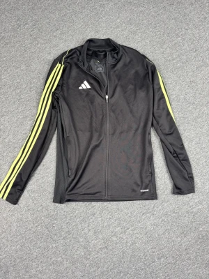 Svart Adidas träningsjacka med gula ränder - Svart träningsjacka från Adidas med hel dragkedja och klassiska gula ränder längs ärmarna. Jackan har lång ärm, ståkrage och är tillverkad i ett lätt, ventilerande material. Perfekt för träning eller sportiga outfits.