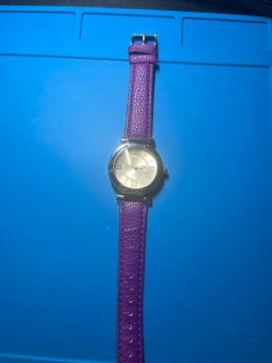 Lila klocka med silverfärgat ur - Stilren klocka med lila armband i skinnimitation och ett runt, silverfärgat ur med romerska siffror. Klassisk design som passar till många olika looks. Armbandet har justerbara hål behövs nya batterier 