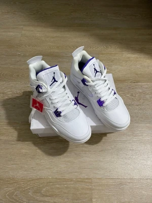 Air Jordan 4 White Purple sneakers - Säljer ett par Air Jordan 4 sneakers i vitt läder med lila detaljer på snörning, plös och sula. Klassisk siluett med meshpaneler och Jumpman-logga på plösen. Perfekta för dig som vill sticka ut med stilrena och sportiga sneakers.