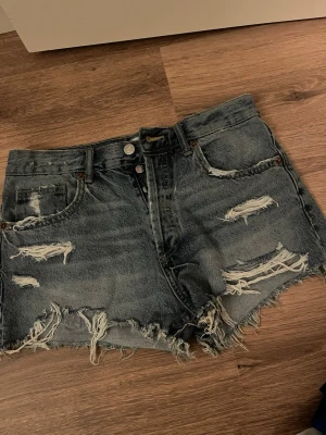 Blå jeansshorts - Säljer ett par blå shorts med slitna detaljer och fransig kant. Shortsen har låg midja.