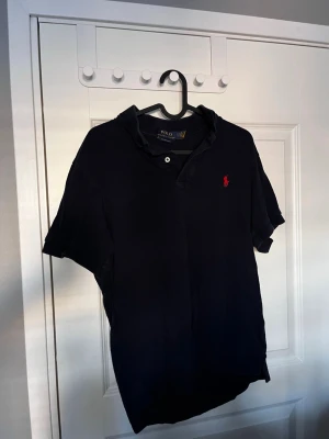 Marinblå pikétröja från Ralph Lauren - Klassisk marinblå pikétröja från Polo Ralph Lauren med röd broderad logga på bröstet. Slim fit-modell med korta ärmar, krage och knappar vid halsen. Tillverkad i mjuk bomull som känns skön mot huden.
