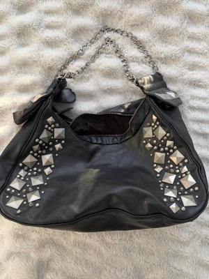 Svart väska med nitar och kedjehandtag - Cool svart handväska i mjukt konstläder med silverfärgade nitar i olika former på framsidan. Väskan har dubbla kedjehandtag och två stora fack upptill. Köpt second hand men har inte sjölv haft användning för den. Funkar som skolväska!