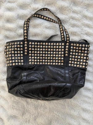 Svart väska med nitar - Cool svart väska i glansigt konstläder med massor av  nitar på ovansidan och handtagen. Väskan har dubbla handtag och rymlig insida, perfekt för att bära med sig allt man behöver. Köpt second hand men har aldrig använt, och finns ingen tidigare tecken på användning. Perfekt som skolväska!
