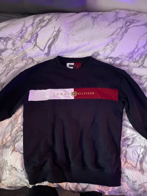 Svart sweatshirt Tommy Hilfiger - Snygg svart sweatshirt från Tommy Hilfiger med klassisk logga och breda ränder i vitt och rött över bröstet. Tröjan har rund halsringning, långa ärmar och liten flaggdetalj på ärmen. Perfekt för en casual och trendig look.
