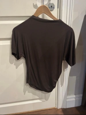 Brun t-shirt med snörning i sidan - Mörkbrun t-shirt med korta ärmar och snygg snörningsdetalj i sidan. T-shirten har en enkel, avslappnad passform och är tillverkad i ett mjukt material som känns skönt mot huden. Perfekt för dig som gillar stilrena basplagg med en twist.